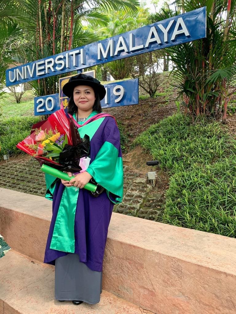 Dr. Norashikin Mohd Shaharuddin in grad robe posing in front of University Malaysa signage