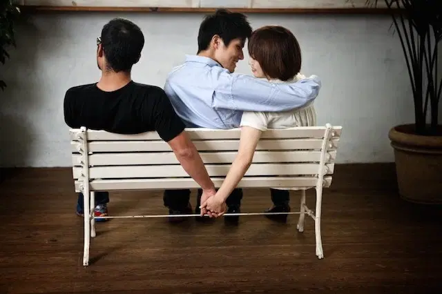 Woman holding a man's hand while she's in a relationship with her lover who sits and wraps his hand around her