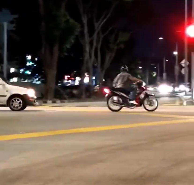 M'sian motorcyclist rides through traffic at night holding gas cylinder with one hand