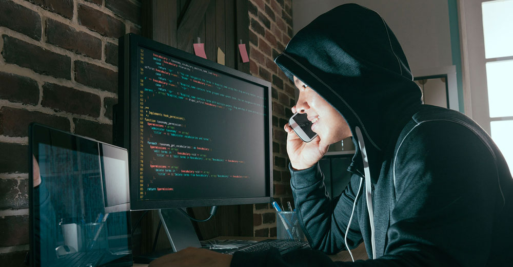 Hacker in black hoodie smiling while calling in front of computer portrayed as a hacker