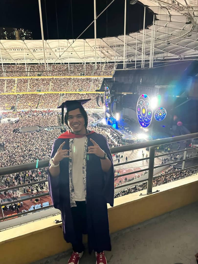Msian graduate wearing his robe at a coldplay concert, taking a picture before the concert starts