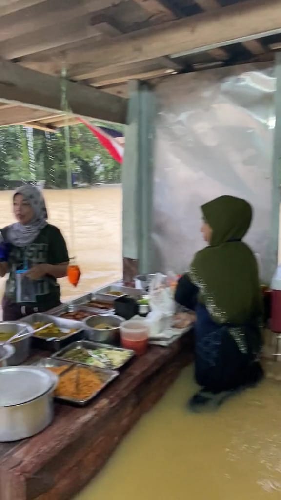 The seller is packing the food requested by her customer despite standing in floodwater rising above her knee level.