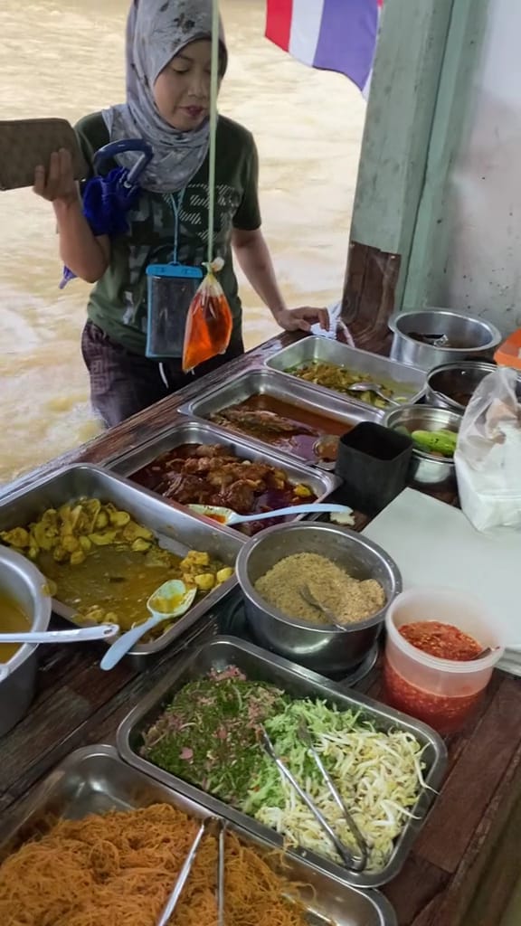 A woman is selling a variety of dishes, including nasi kerabu, noodles, and other side dishes, at the stall in front of her house.